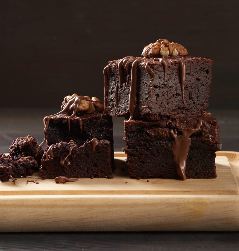 Stack of baked square pieces of chocolate brownie cake on brown wooden cutting board