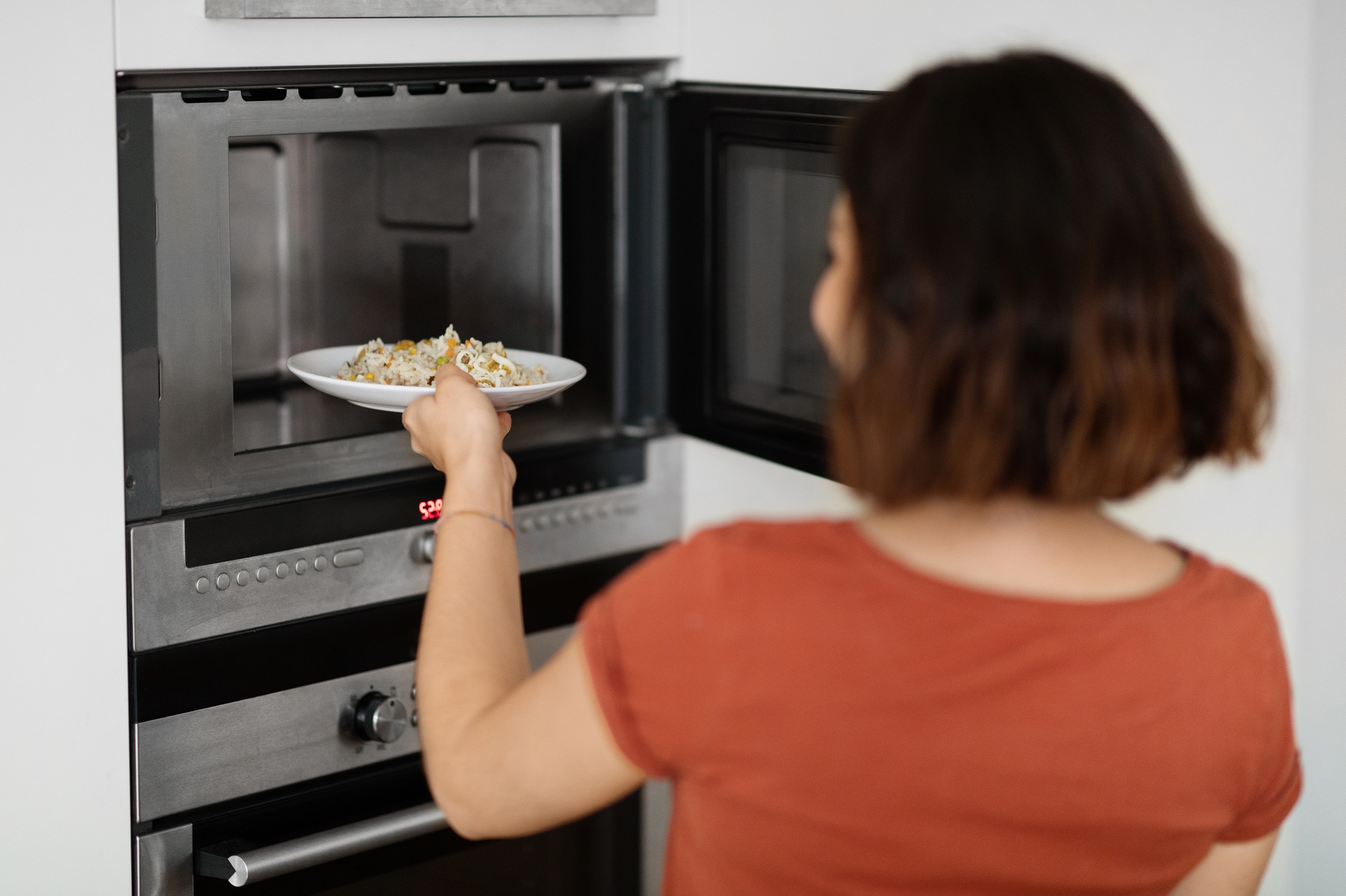 Young Woman Heating Rice With Vegetables Dish In Microwave Oven At Home